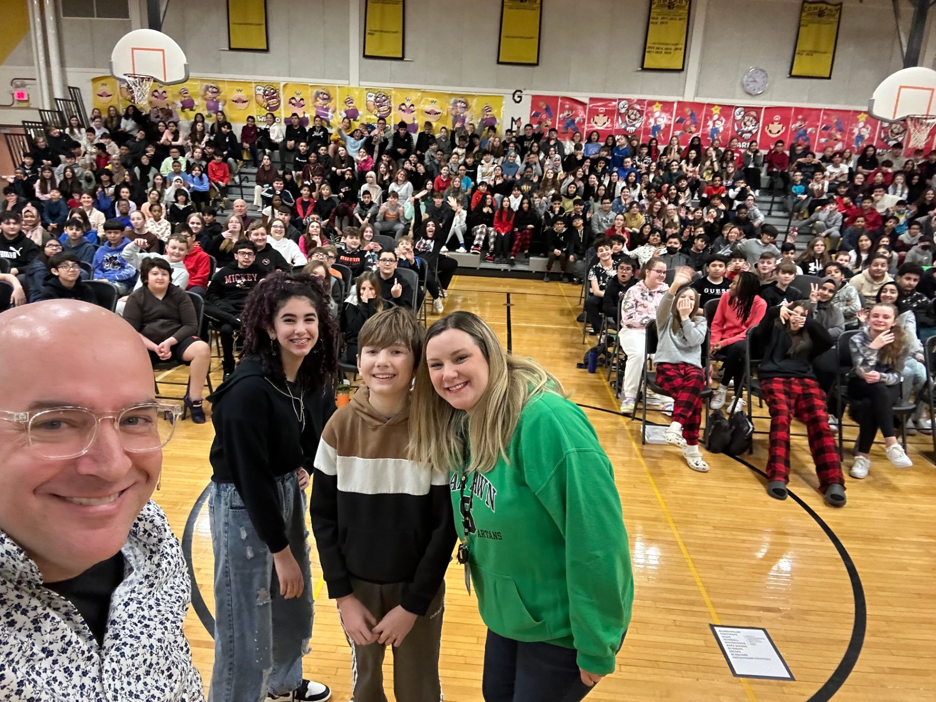 Tim taking a selfie with three student volunteers in front of a packed middle school gym